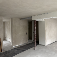 Sicht vom Wohnzimmer in den Eingangsbereich der Wohnung. Innenputz und Estrich sind eingebracht. Eine Stahlstütze steht unter einem verkofferten Stahlträger