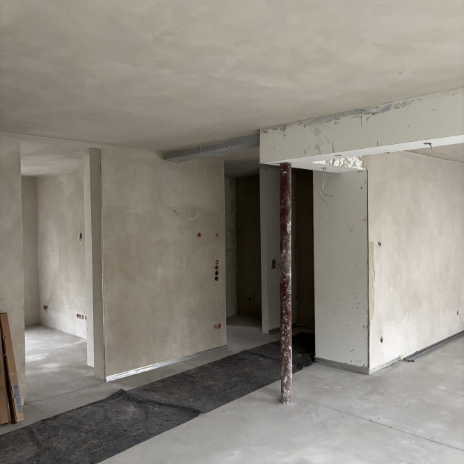 Sicht vom Wohnzimmer in den Eingangsbereich der Wohnung. Innenputz und Estrich sind eingebracht. Eine Stahlstütze steht unter einem verkofferten Stahlträger